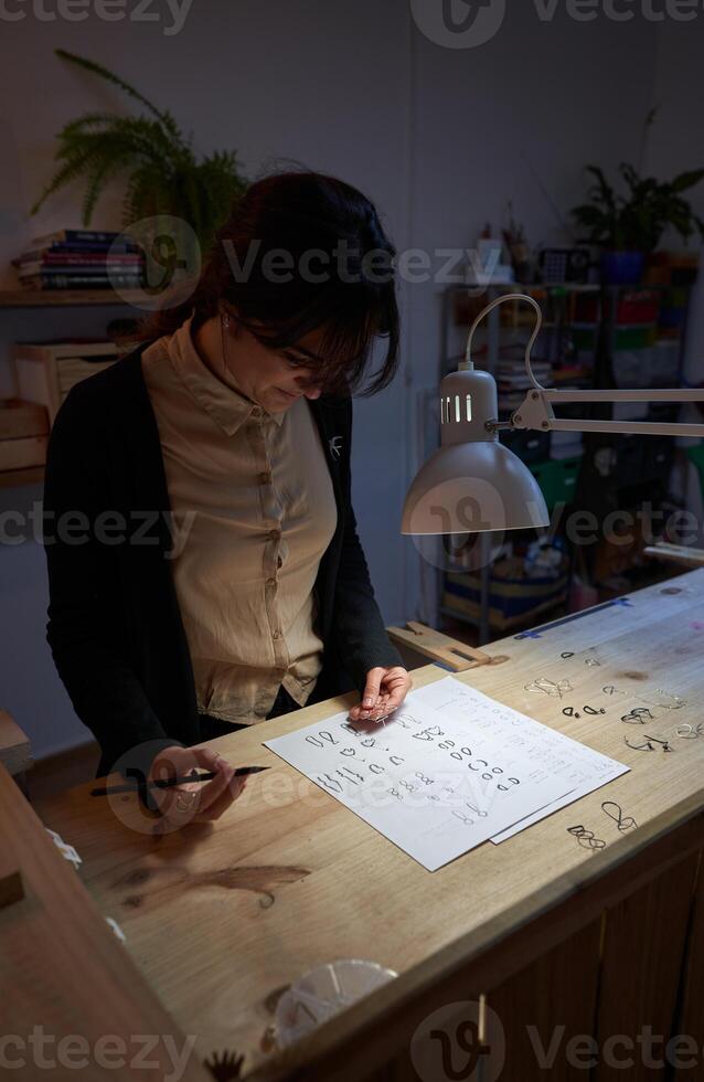 Female goldsmith drawing jewelry pictures at table in dark room photo