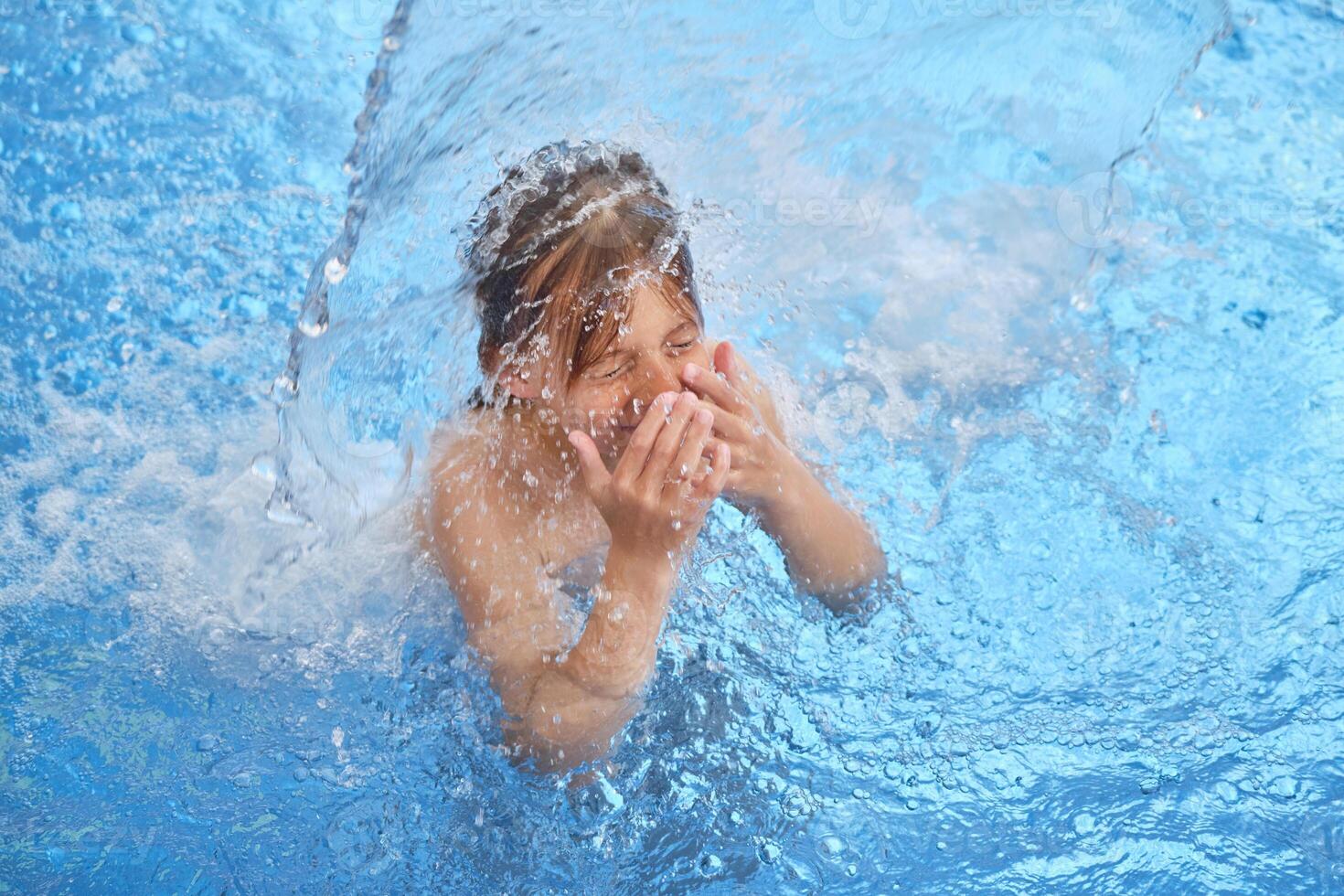 Boy covering nose standing under splashing water in pool photo
