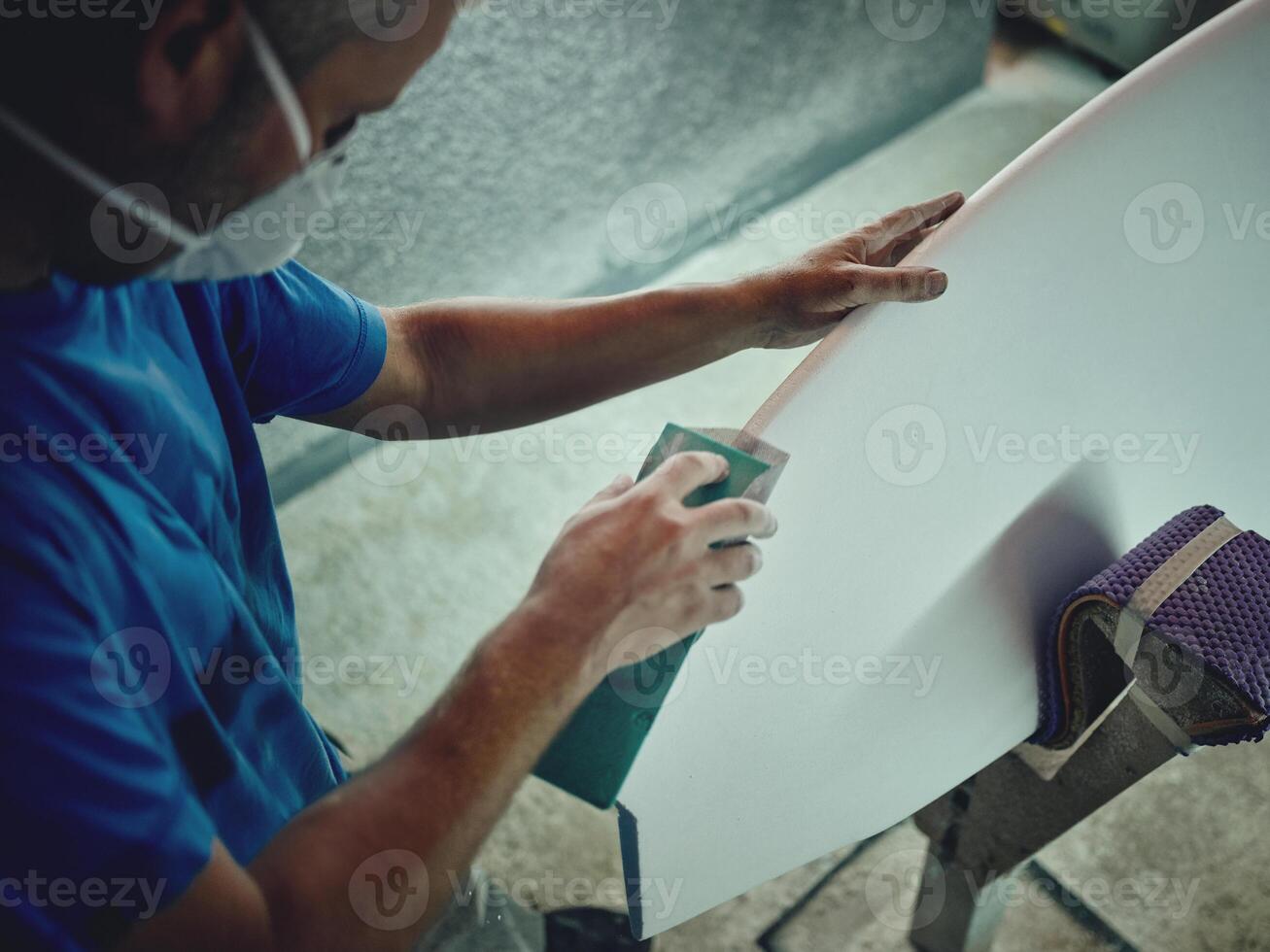 Carpentry man in respirator polishing surfboard with abrasive photo
