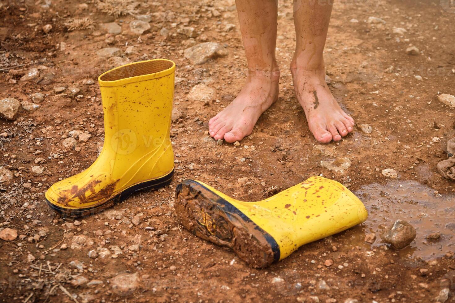 Barefoot kid standing on ground near gumboots photo