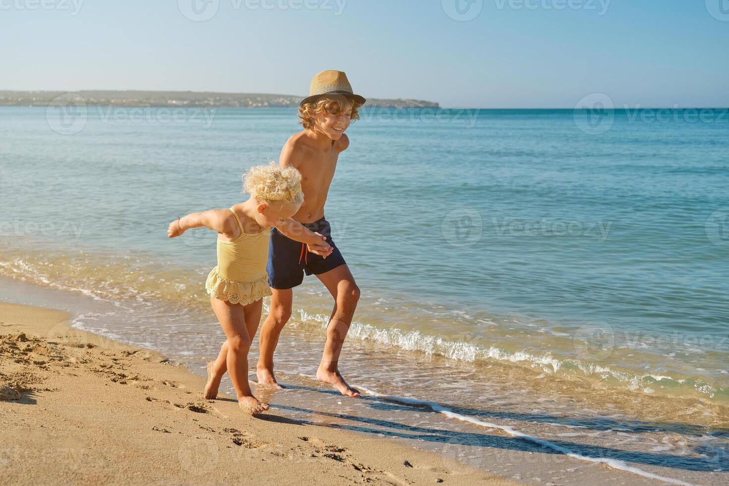 Happy smiling boy holding hand of little girl on beach and runni photo