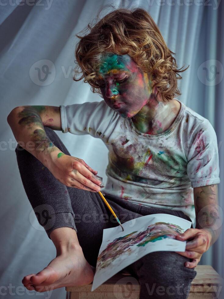 Boy with arm put on knee holding paintbrush and drawing photo