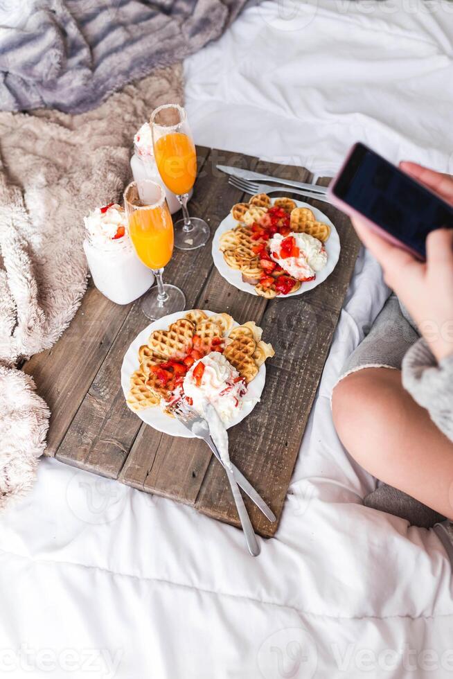 Woman with smartphone near appetizing waffles and tasty drinks on tray on bed photo
