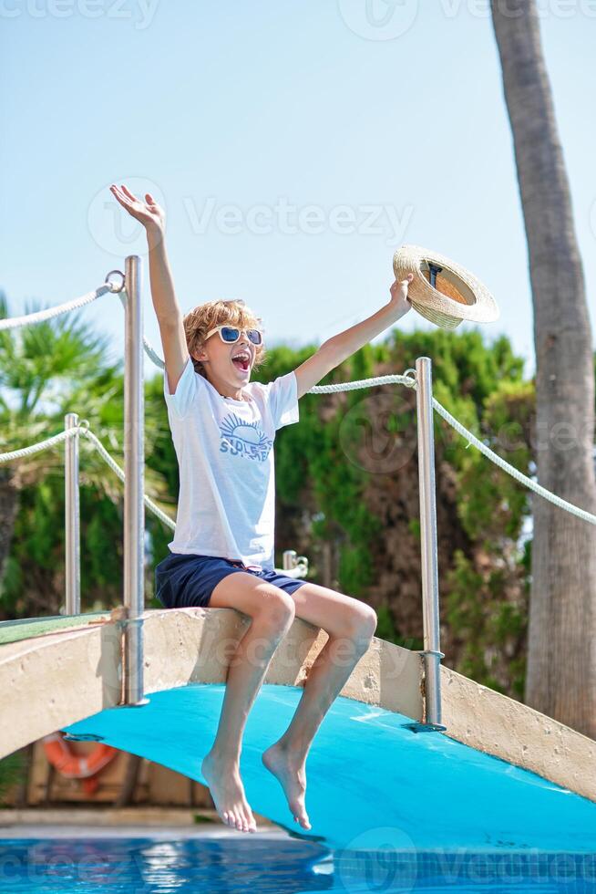 Low angle cheerful boy sitting on footbridge over swimming pool at resort photo