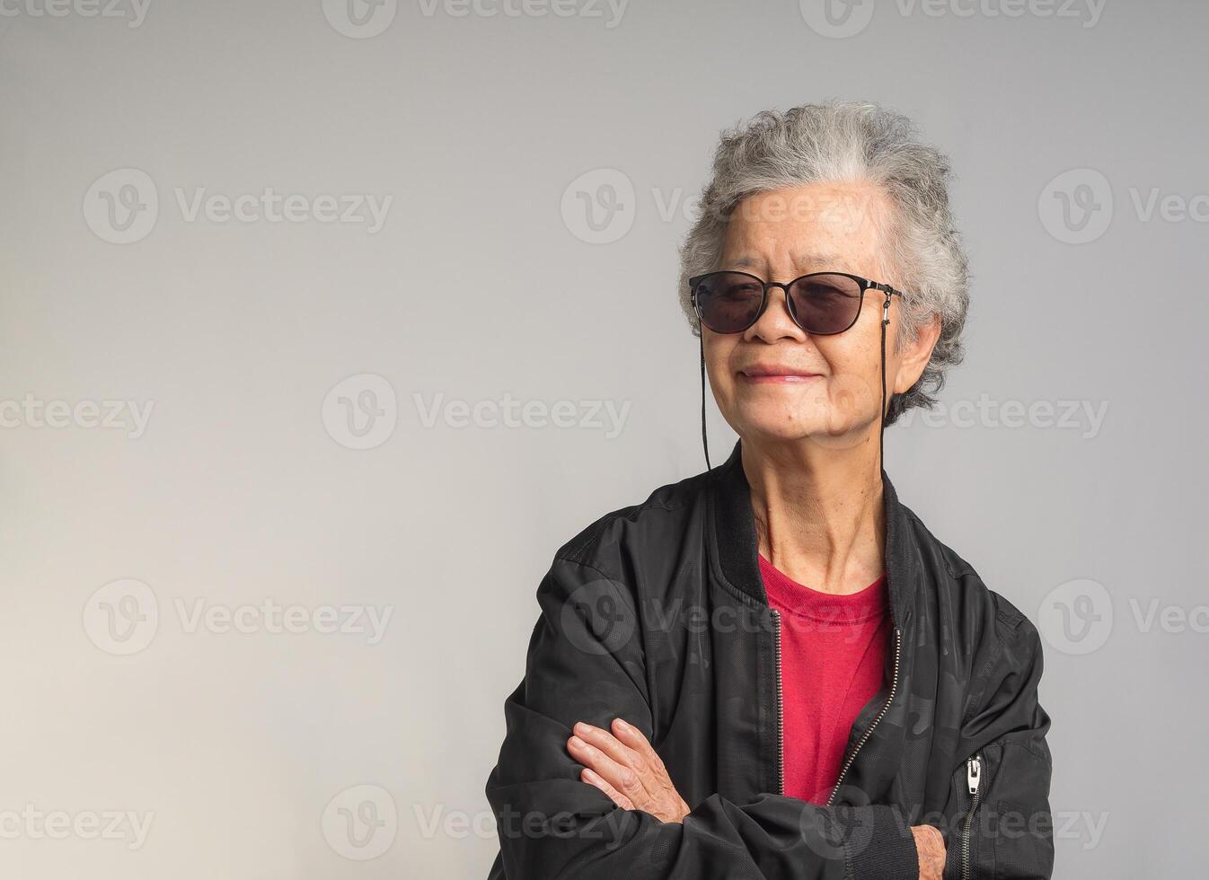 An elderly Asian woman in a black jacket wearing sunglasses while standing with arms crossed on a gray background. photo