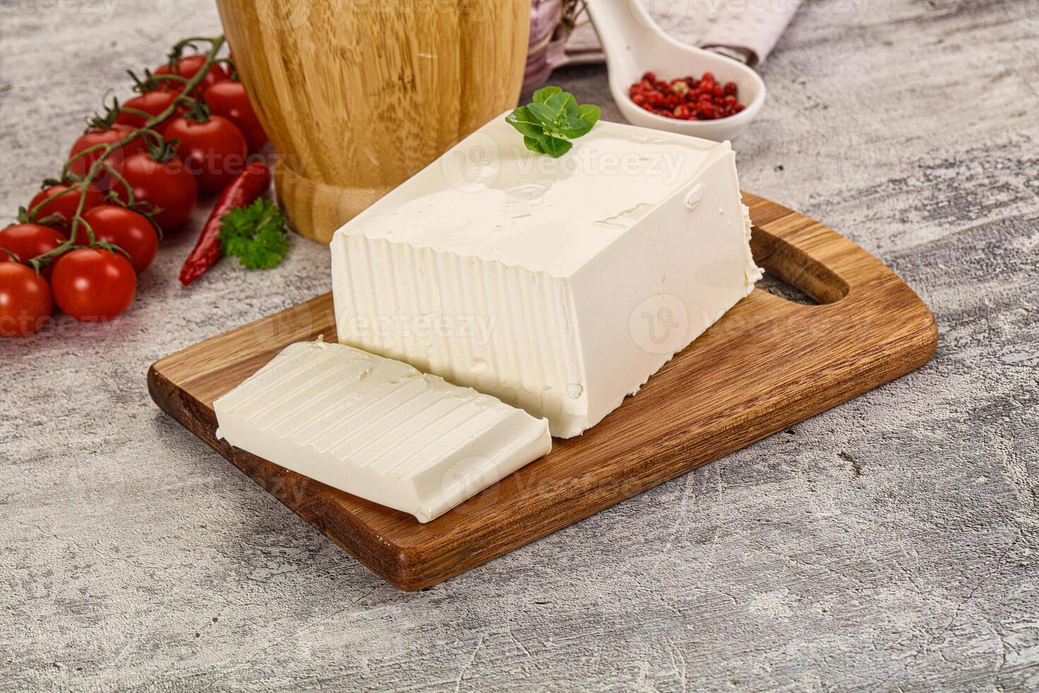 A piece of cheese on a cutting board next to tomatoes and a wooden bowl photo