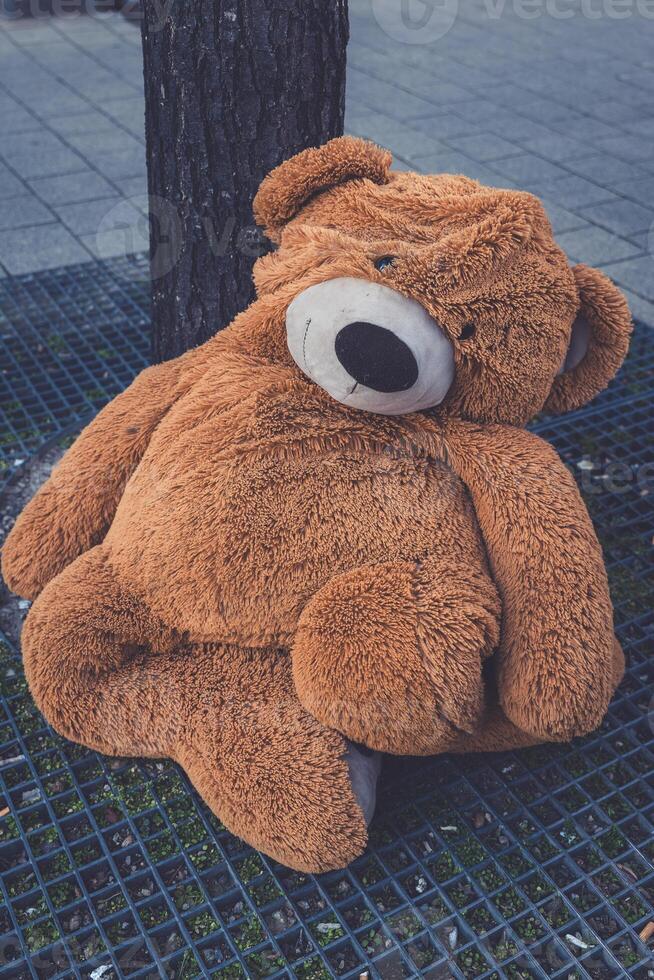 Giant stuffed bear sitting against a tree on a city street during the day, creating a charming urban scene photo
