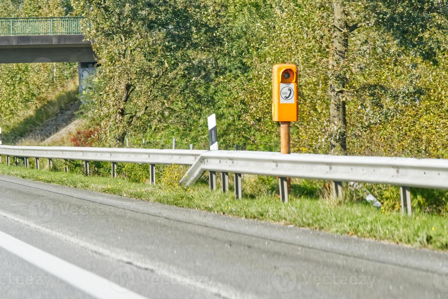 A roadside view featuring a speed camera mounted on an orange pole, with a highway and green trees in the background. The road is clear and well-maintained, indicating a rural or suburban area. photo