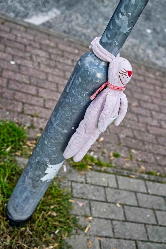 A lost pink stuffed bunny doll tied to a metal pole on a sidewalk, with a grassy area nearby. The doll has a stitched face and is wearing a red scarf. photo