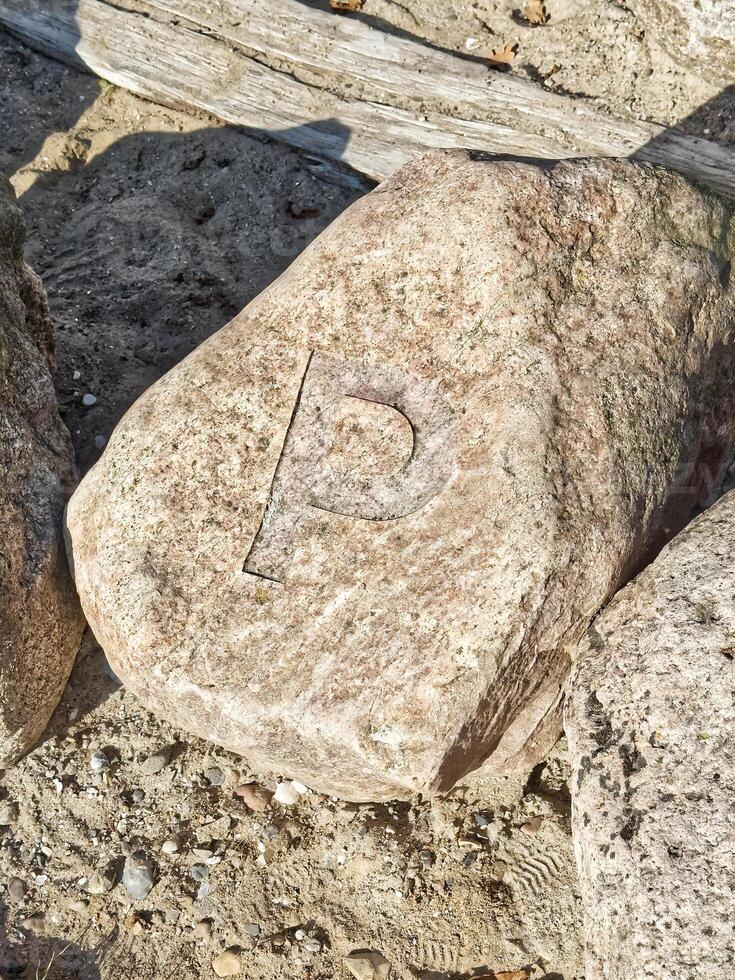 A large, weathered rock with a carved letter P on its surface, surrounded by sandy terrain and other stones. photo
