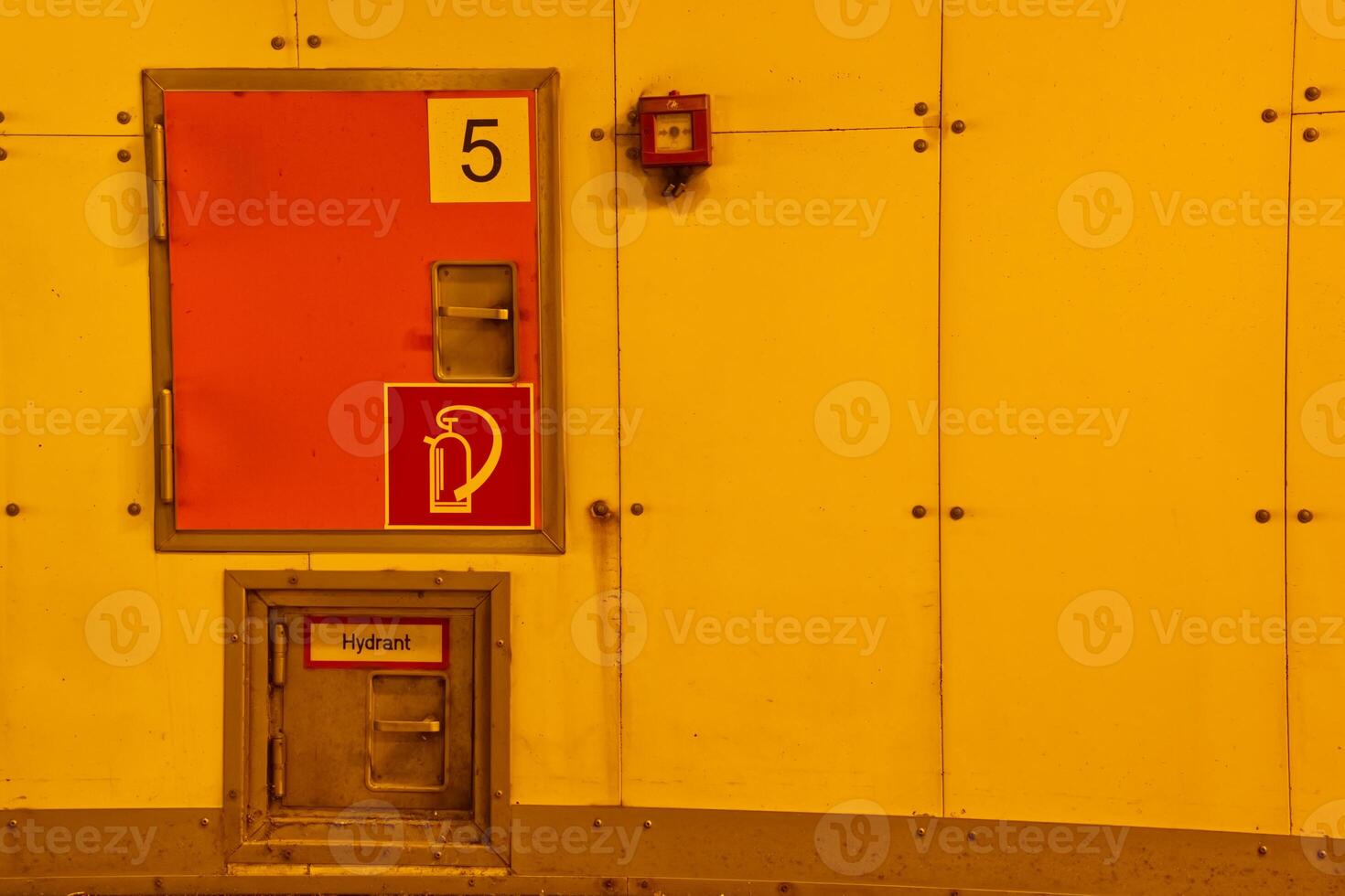 A bright yellow wall featuring a fire hydrant access door and a fire alarm. The door is marked with the number 5 and includes a fire hydrant symbol. The overall color scheme is warm and industrial. photo