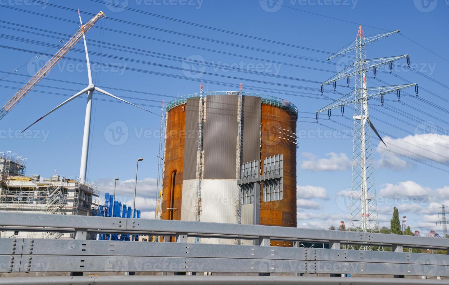 A large industrial structure with a cylindrical shape, surrounded by wind turbines and power lines. The scene captures a mix of renewable energy and industrial development under a clear blue sky. photo