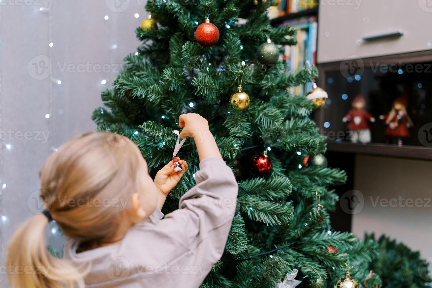 Little girl hangs a snowman figurine on a colorful Christmas tree in the room. Back view photo