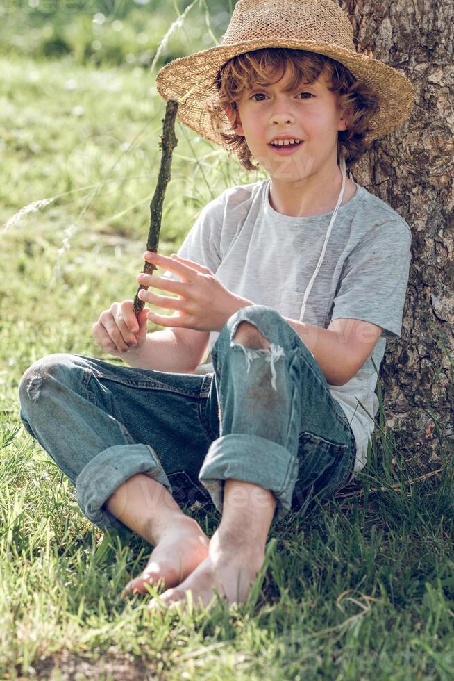 Cheerful boy in wicker hat and with stick under tree photo