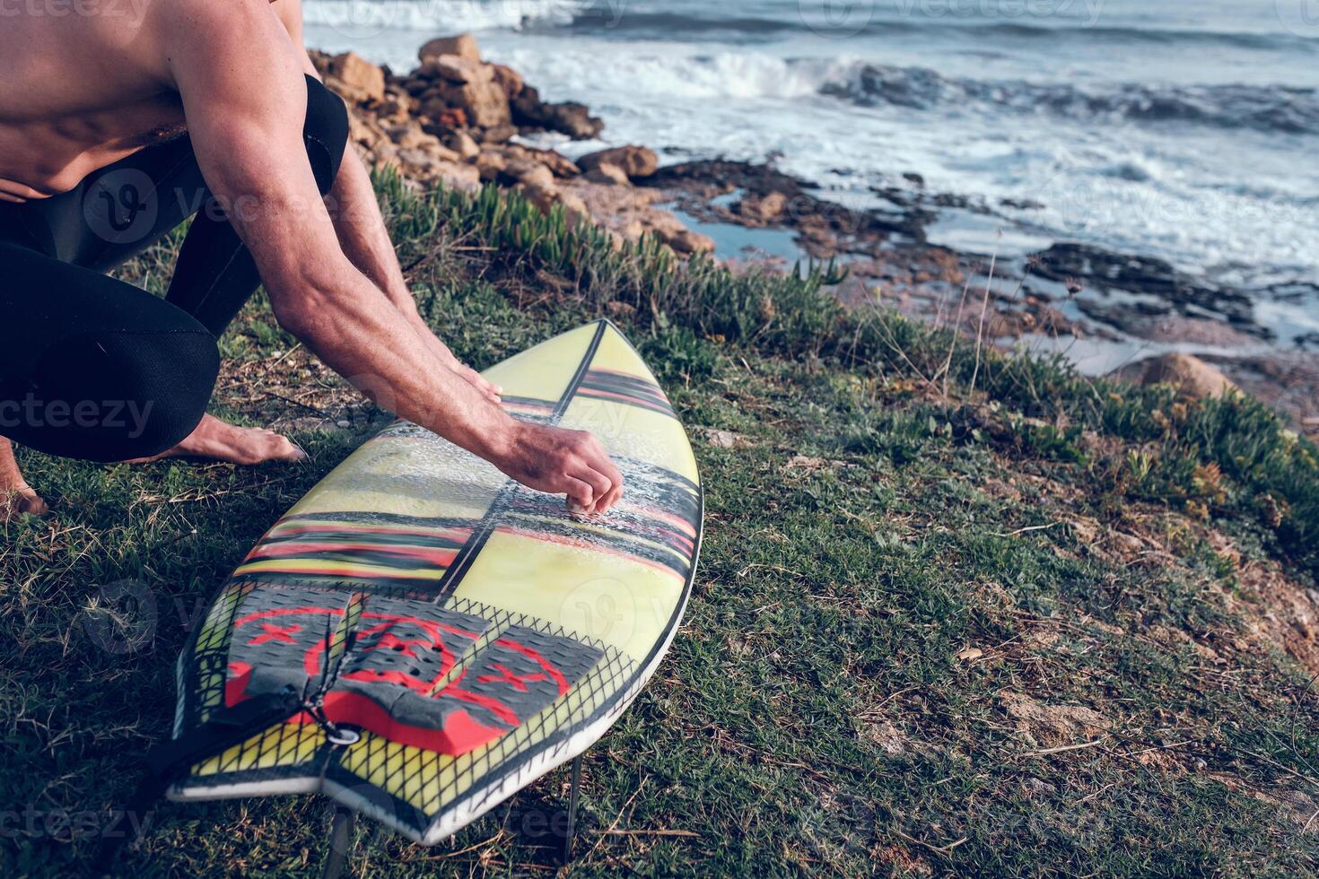 Crop male surfer rubbing surfboard with wax on rocky seashore photo