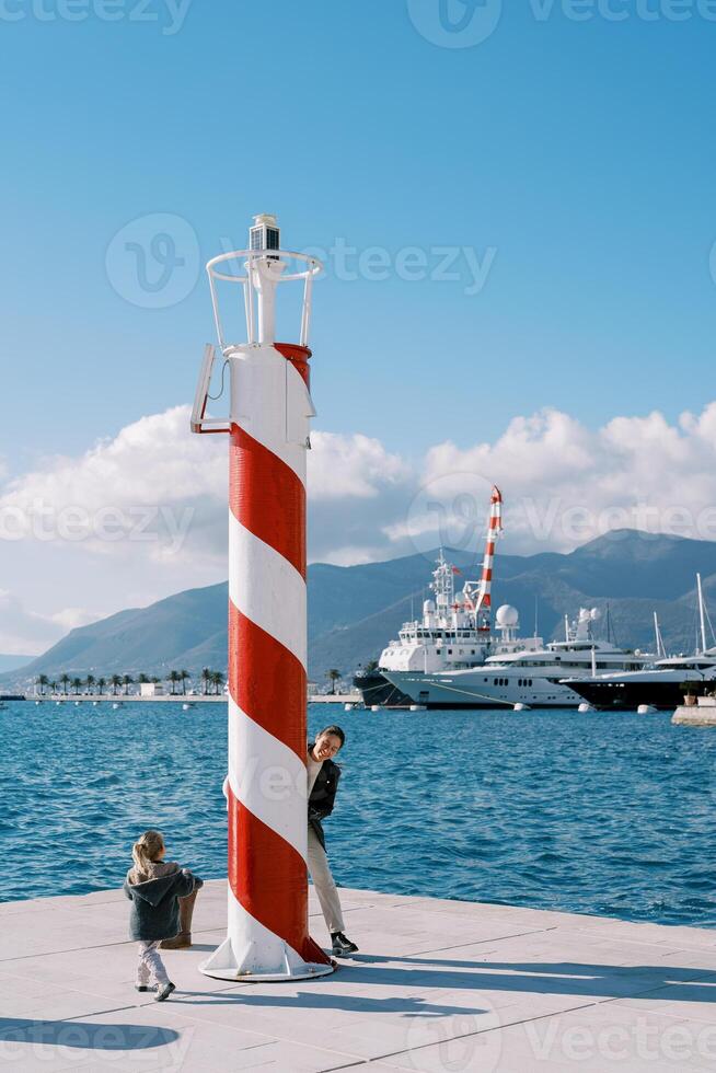 Smiling mother playing hide and seek with little girl near lighthouse on pier photo
