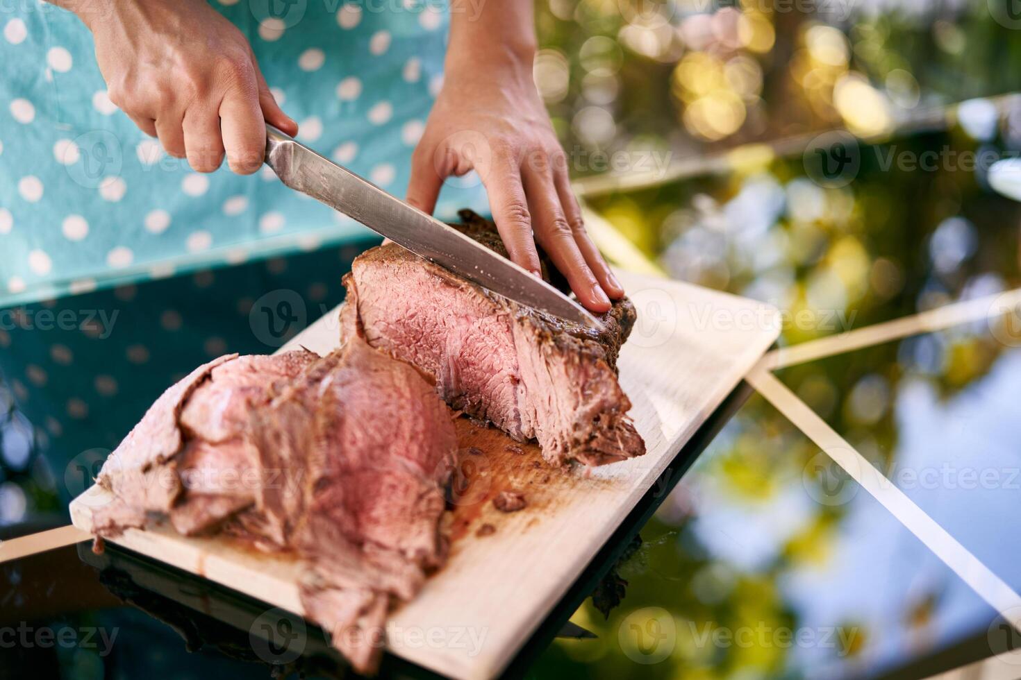 Housewife with a large knife cuts slices of juicy roast beef on a cutting board on a mirror table photo
