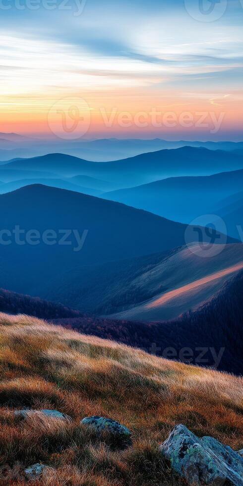 breathtaking landscape showcasing rolling mountains at sunset, with layers of blue and orange hues creating serene atmosphere. foreground features grassy hills and rocky outcrops photo