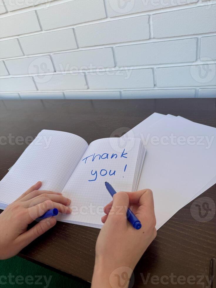 A close-up view of a young woman's right hand writing Thank You in blue marker on a notebook, against the background of a dark wooden table with white brick wall photo