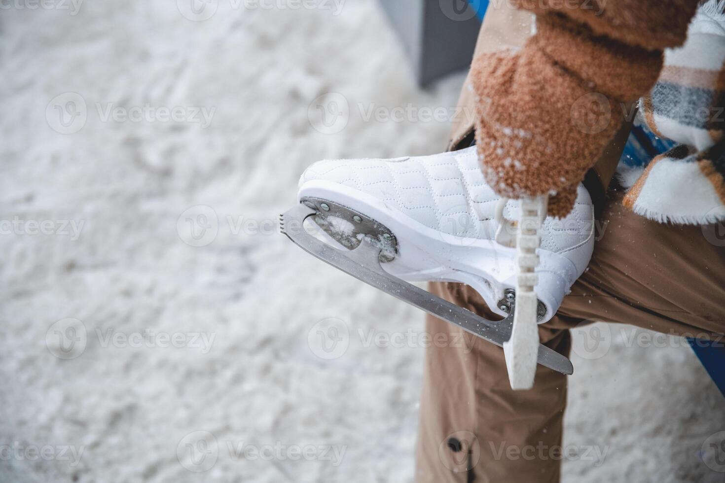 Woman wearing brown gloves and beige pants is putting on white ice skates while sitting on a blue bench in an outdoor ice rink, getting ready for a fun day of skating photo
