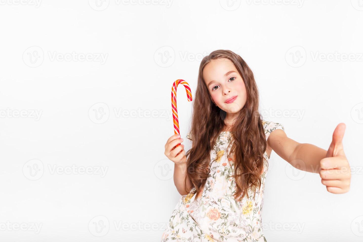 Happy, smiling cute little girl eating cristmas candy cane. Saying Ok. Posing against a white wall. photo