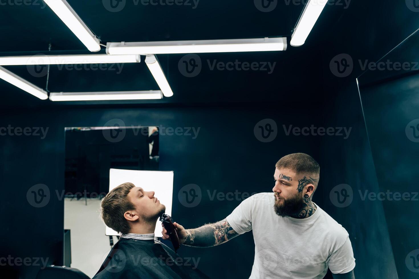 Tattooed Barber trimming bearded man with shaving machine in barbershop. Hairstyling process. Hairstylist cutting the beard of a bearded male. photo