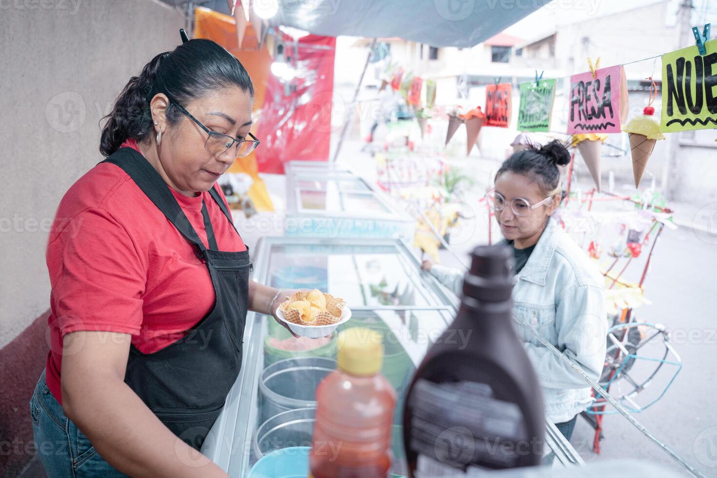 An adult Hispanic woman is preparing a vanilla ice cream for a young woman client in a street market stall photo