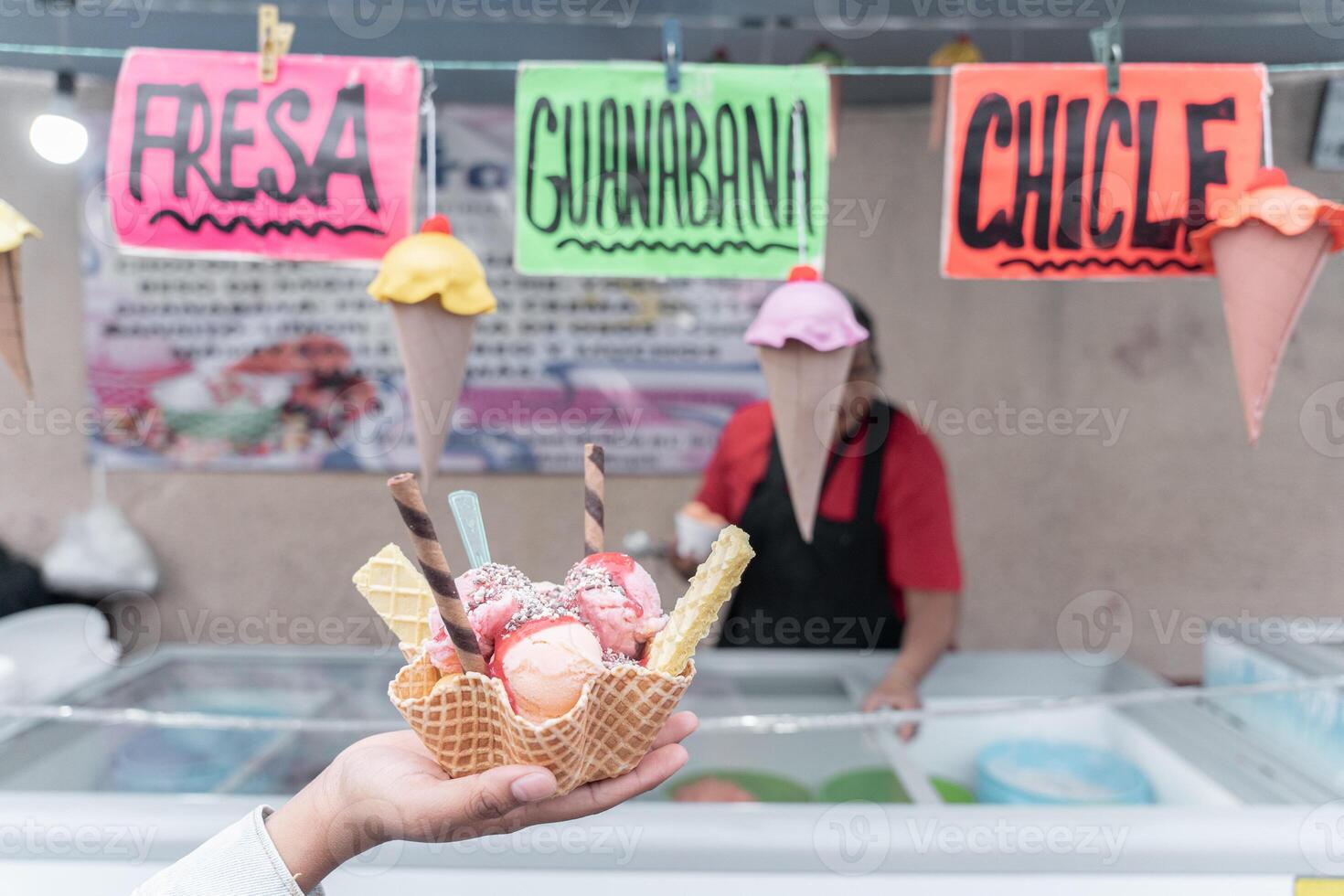 A hand is holding a traditional Mexican decorated ice cream in front of a street market stall with flavor signs photo