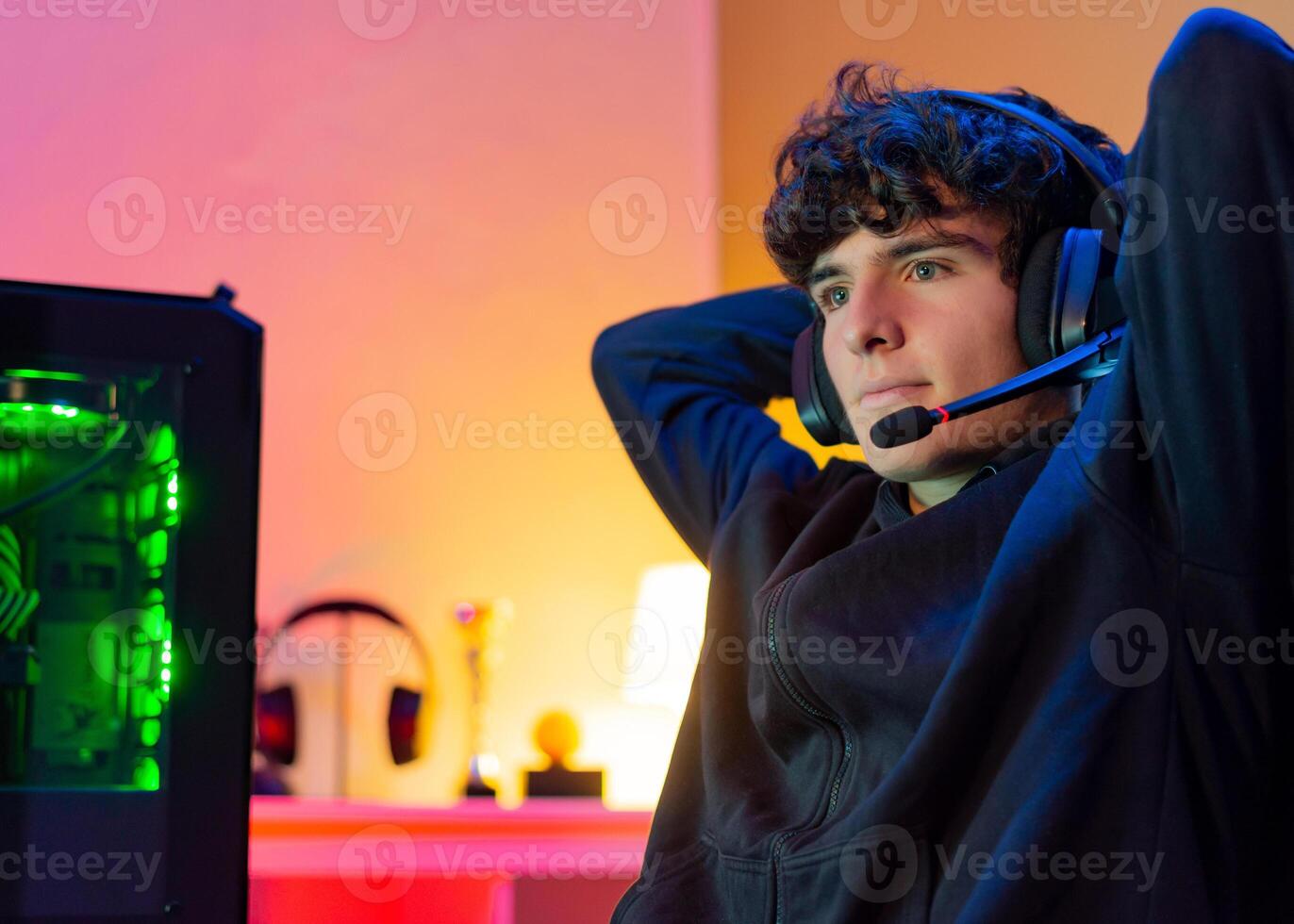Young Caucasian teenager gamer with headphones leans back in chair with arms behind head with satisfied face. photo