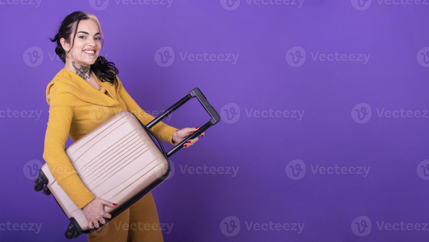 Attractive happy woman traveler holding her suitcase like a guitar against purple background. Copy space. photo