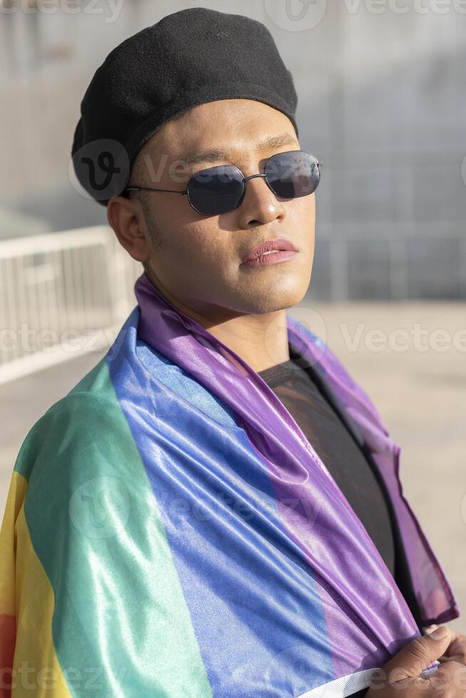 Gay man with make up on under LGBT rainbow flag while looking at camera. Concept of Pride. Pride month. photo