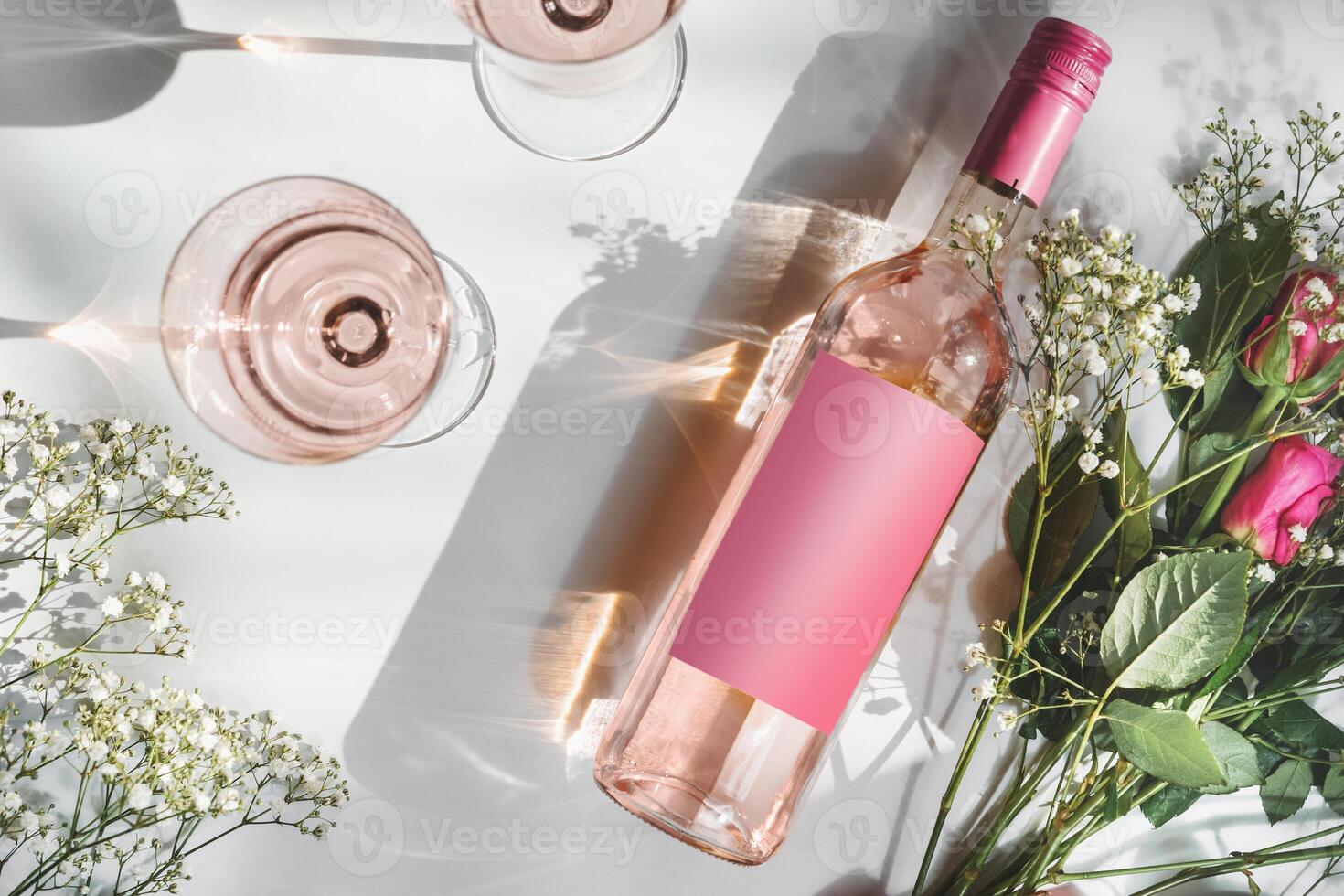 Rose wine bottle with empty pink mock up label, two wine glasses and romantic flower bouquet on white background with sunlight and shadow. Modern wine concept. Top view photo