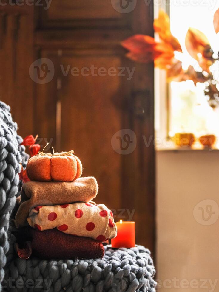 Autumn still life with folded cozy sweaters, wool blanket, pumpkin, candle at window background with sunlight. Warm clothes and seasonal interior decoration at home. Front view photo