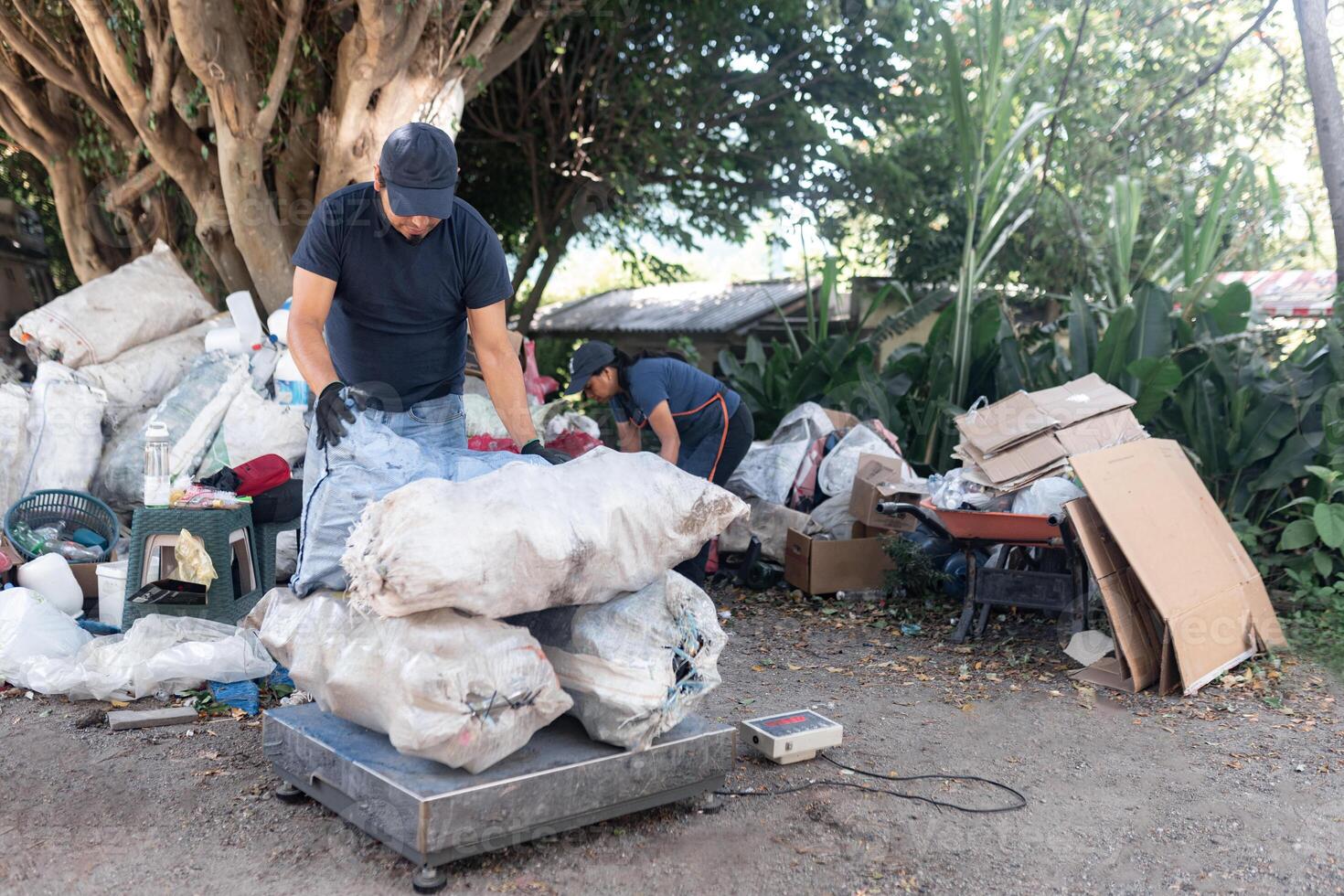 An adult Hispanic man weighs a pile of sacks of reclaimed materials, supported by a woman in the background photo