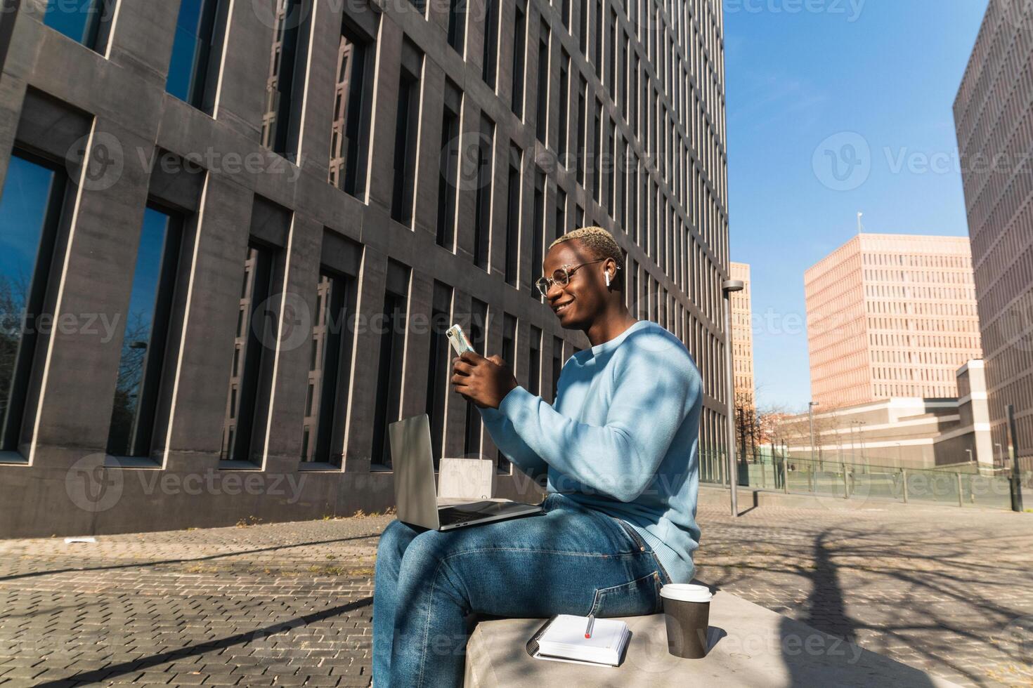 Self employed young ethnic guy using smartphone during work on netbook on street photo