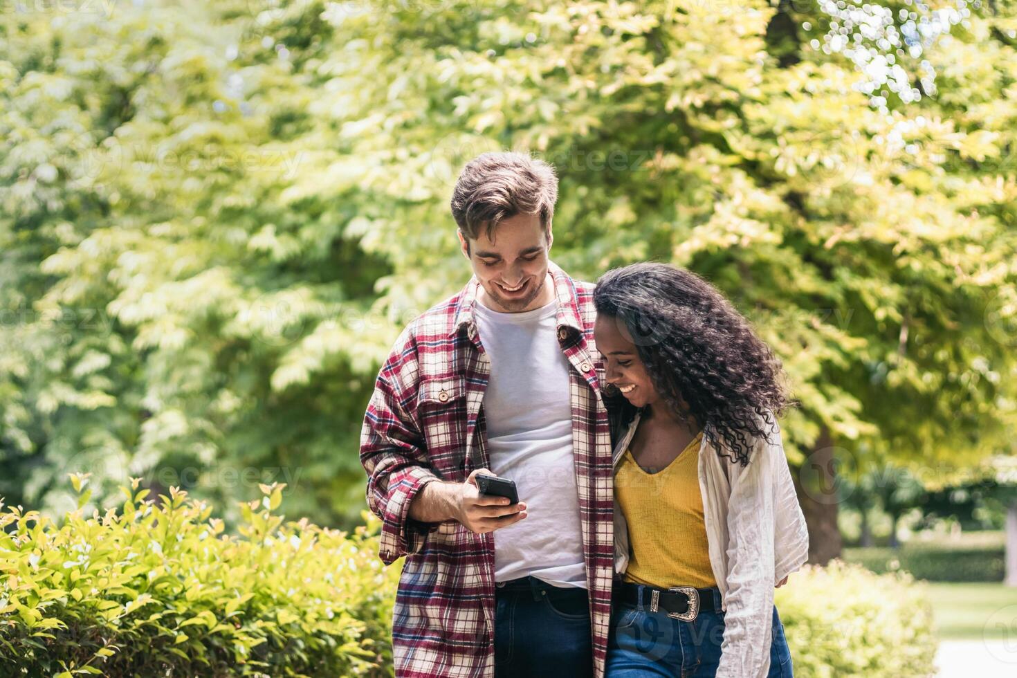 Happy multiracial couple taking a walk in the park using smartphone photo
