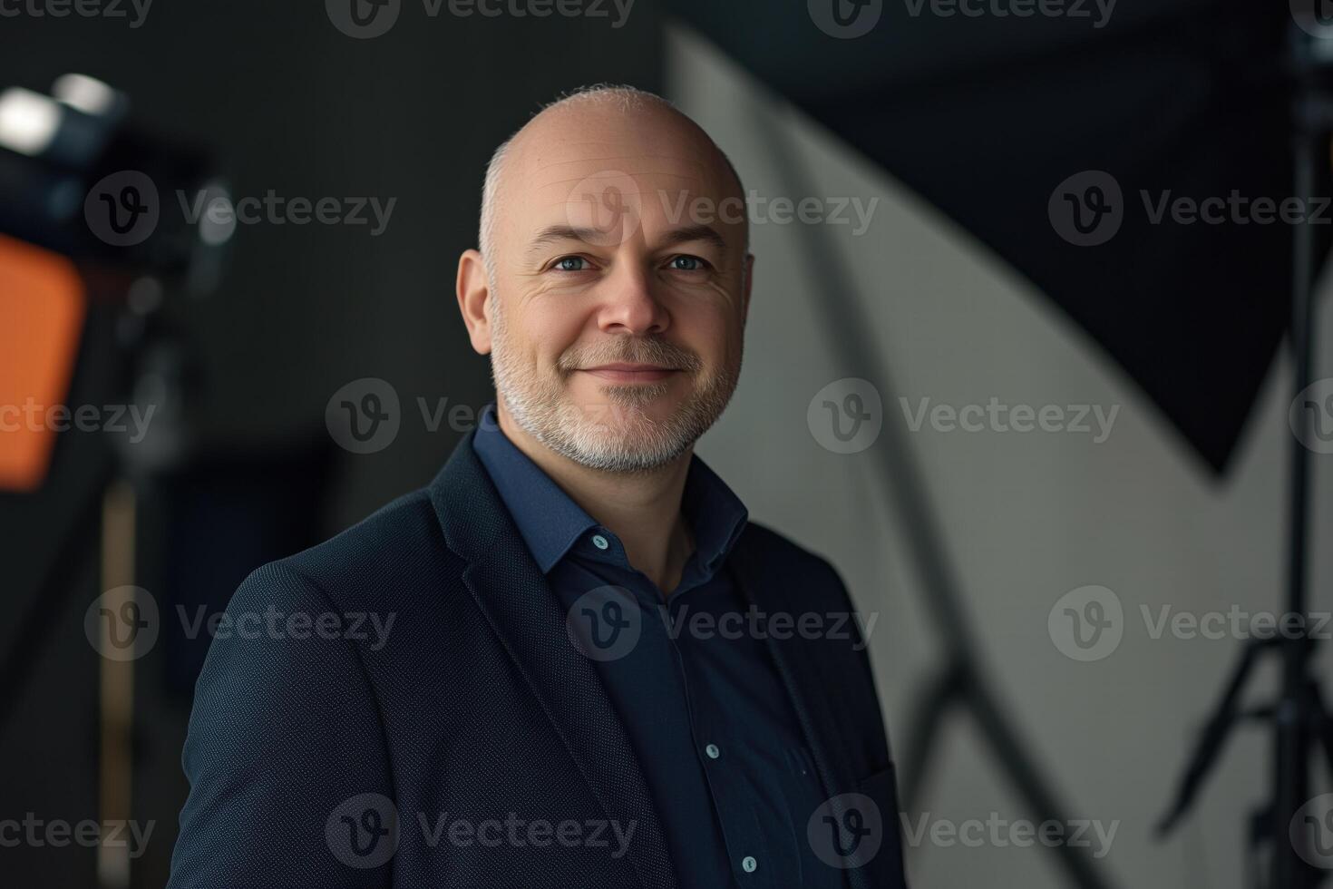 A confident middle-aged sales manager man with a short beard, wearing a dark suit and blue shirt, smiles warmly at the camera. The background is softly blurred, with studio lighting equipment visible. photo