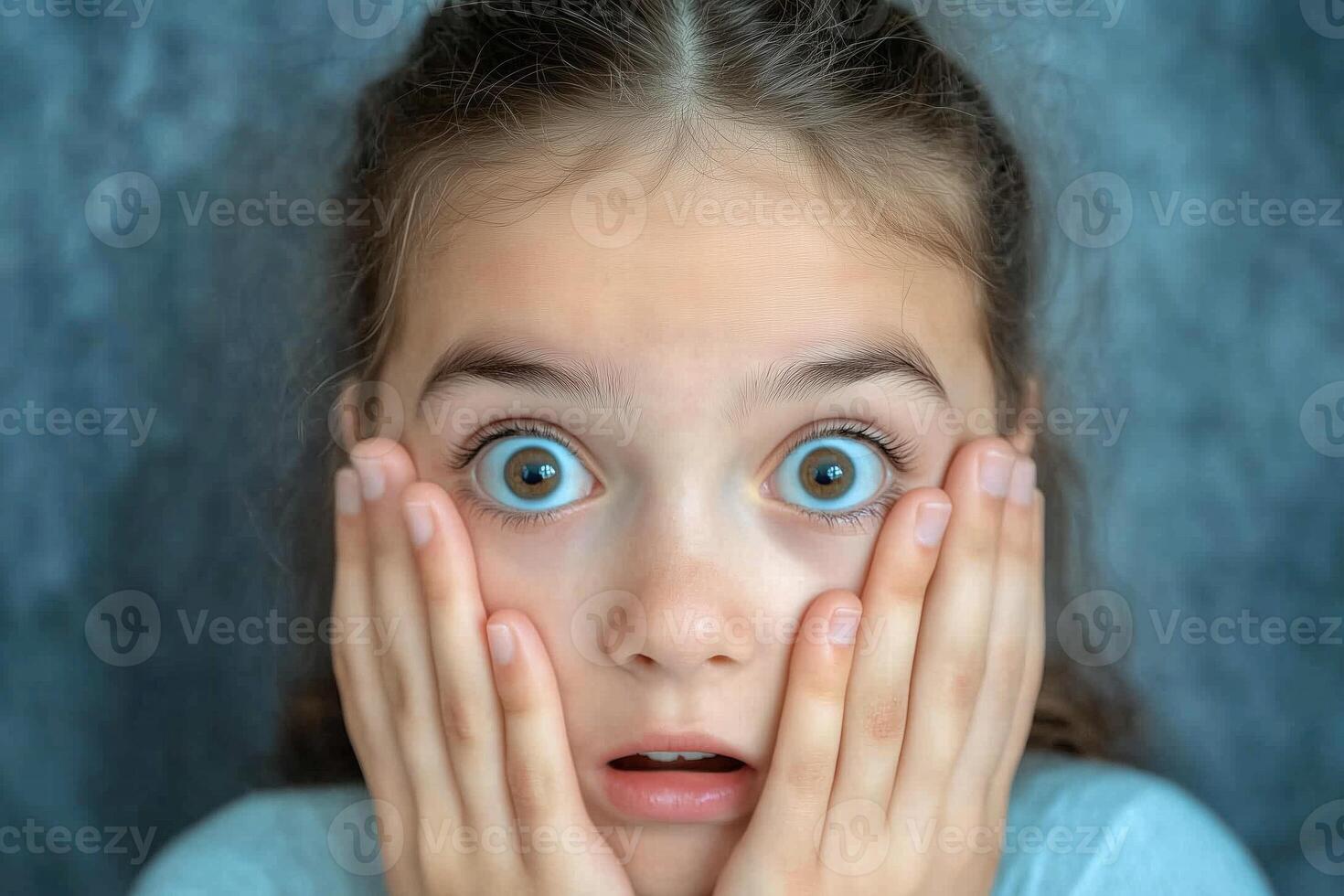 A young girl with big blue eyes is holding her hands to her face photo