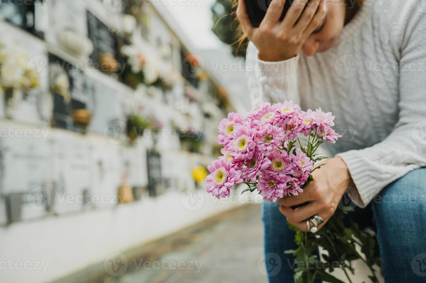 Grieving woman touching forehead while sitting near columbarium wall in cemetery photo
