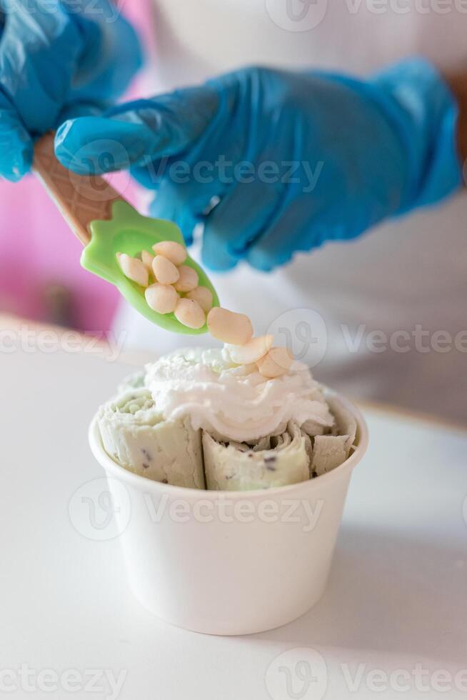 A worker is decorating a rolled ice cream tub with yogurt bubble toppings with a spoon photo