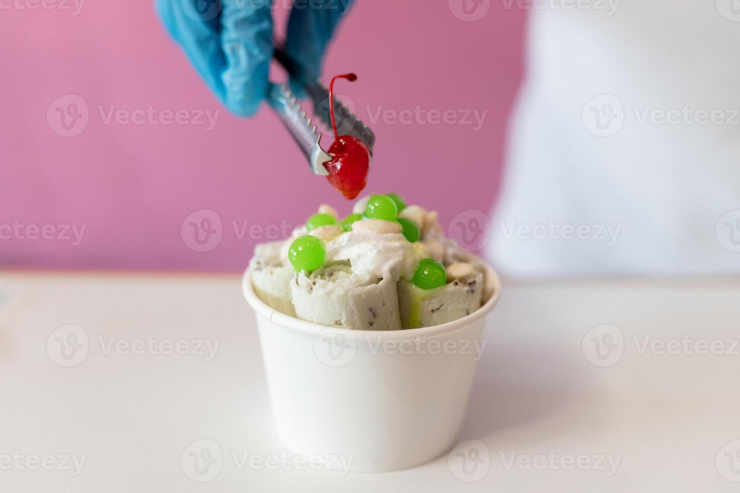 A worker is decorating a rolled ice cream tub with a cherry on top using tongs photo