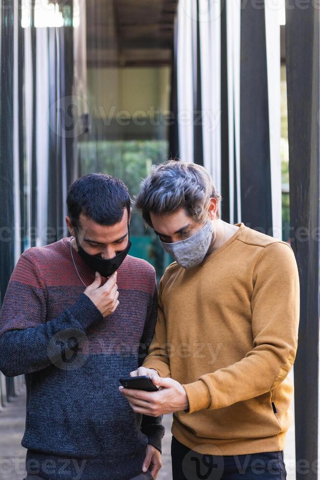Smiling men in masks browsing cellphone together on street photo