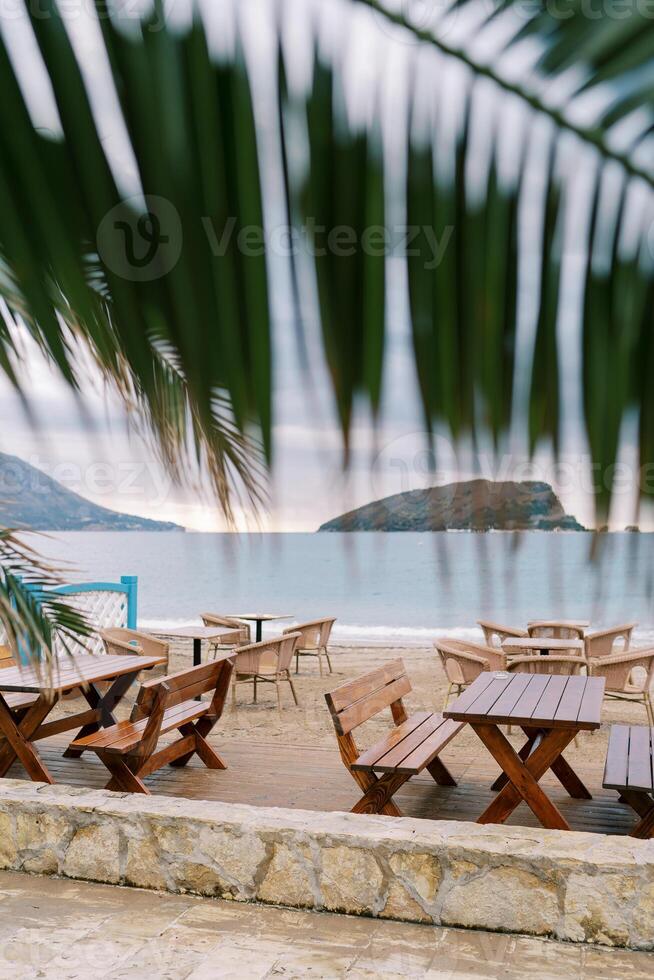 View from behind a green palm leaf on wooden tables and chairs standing on the beach by the sea photo