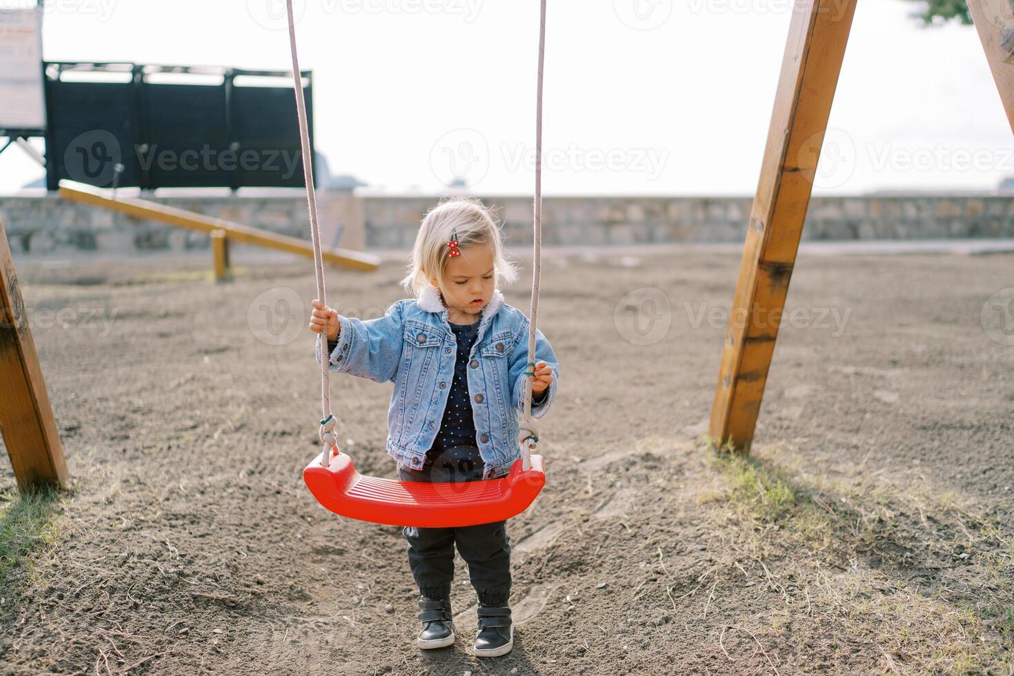 Little girl stands near the swing on the playground holding on to the ropes photo