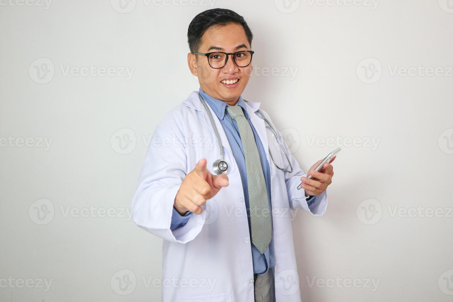 Smiling Asian male doctor pointing finger at camera. Man standing and looking at camera. Medical product concept. Isolated front view on white background. photo