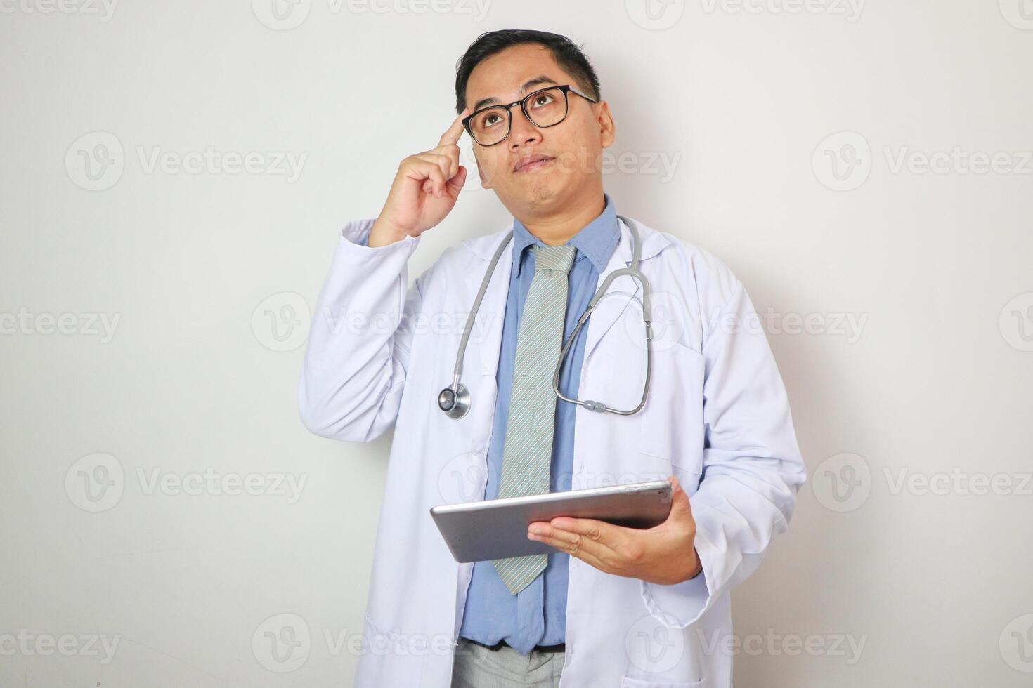 Asian man doctor thinking gesture uses digital tablet in modern medical office. Focused expression indicates consideration and analysis. Concept of technology in healthcare. photo