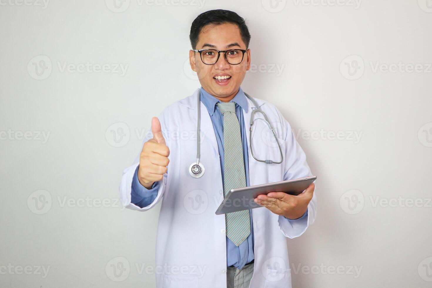 A handsome, smiling Asian male doctor in a white gown uniform showing thumb up while using digital tablet, reading medical cases, standing against on a white studio background. photo