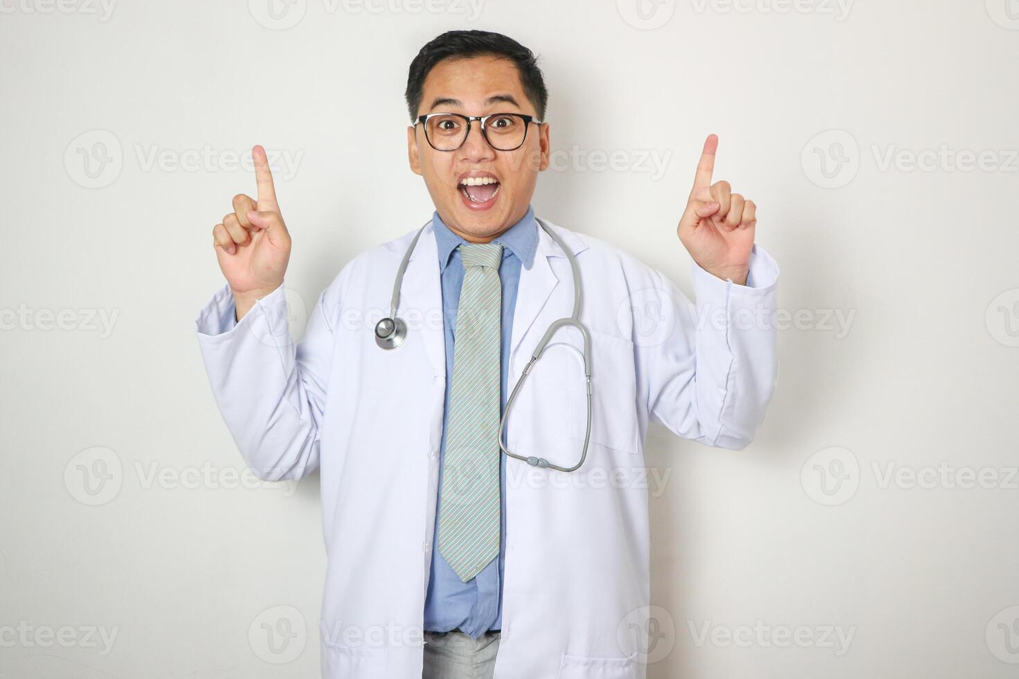 Surprised glad attractive mature asian doctor in white coat, glasses with stethoscope showing finger up on empty space isolated on white background. Therapist got idea, treatment, great offer and ad photo
