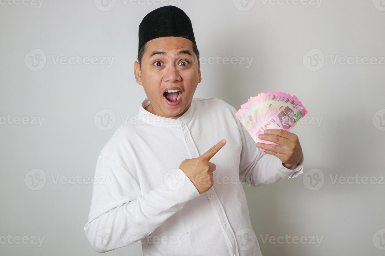 Smiling young Asian Muslim man pointing finger at cash money rupiah banknotes isolated on white background. People religious Islamic lifestyle concept photo