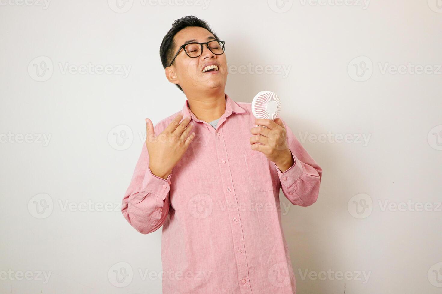 Asian man, dressed in a beanie hat and casual shirt, revels in the airflow and refreshing cool wind from a handheld portable mini fan to beat the summer heat, while standing against a white background photo